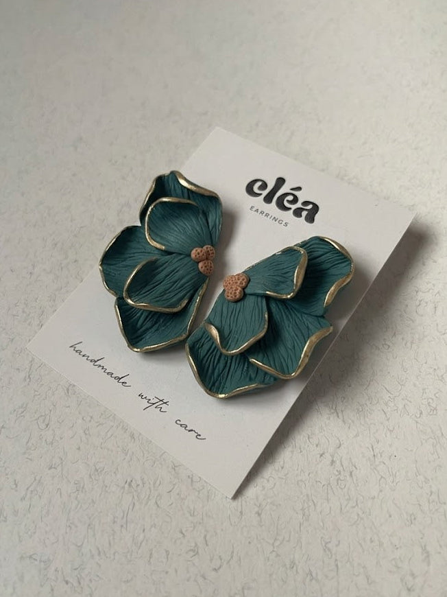 Teal flower-shaped earrings on a card with 'cleá' branding on a light gray background