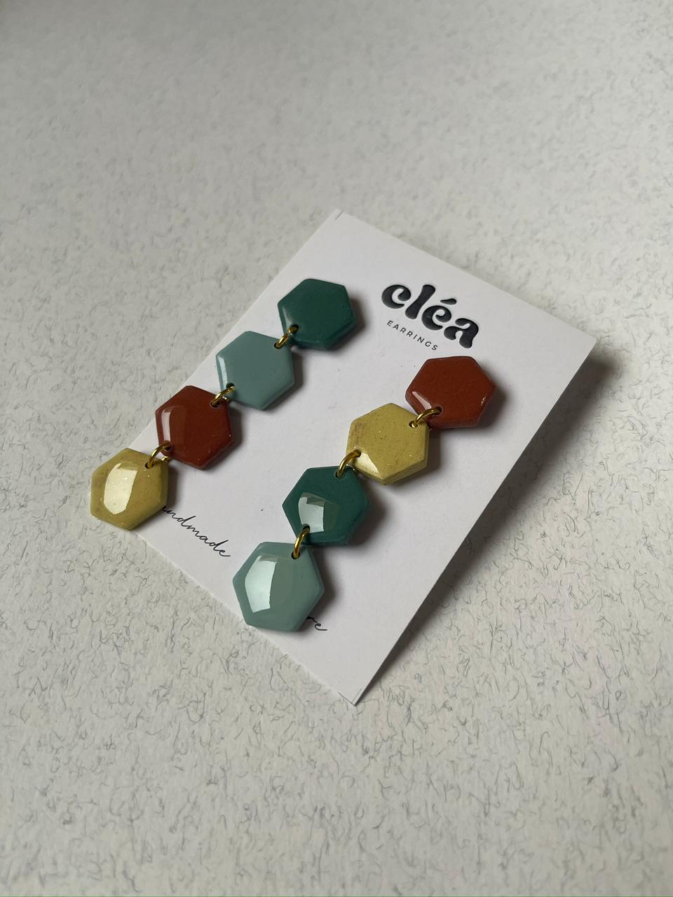 Colorful hexagonal earrings handmade earth tone on a 'cléa' branded card against a wooden background.