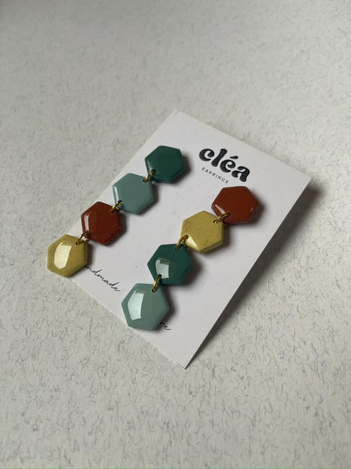 Colorful hexagonal earrings handmade earth tone on a 'cléa' branded card against a wooden background.