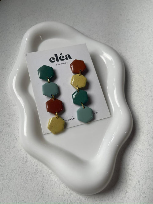 Colorful geometric earrings on a 'Cleá' branded card on a white dish.