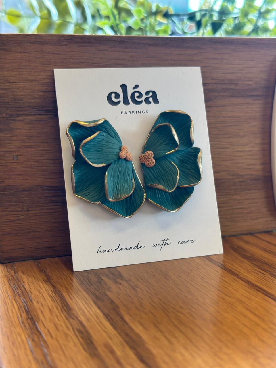 Teal flower-shaped earrings on a Clea branded card, displayed on a wooden surface with greenery in the background.